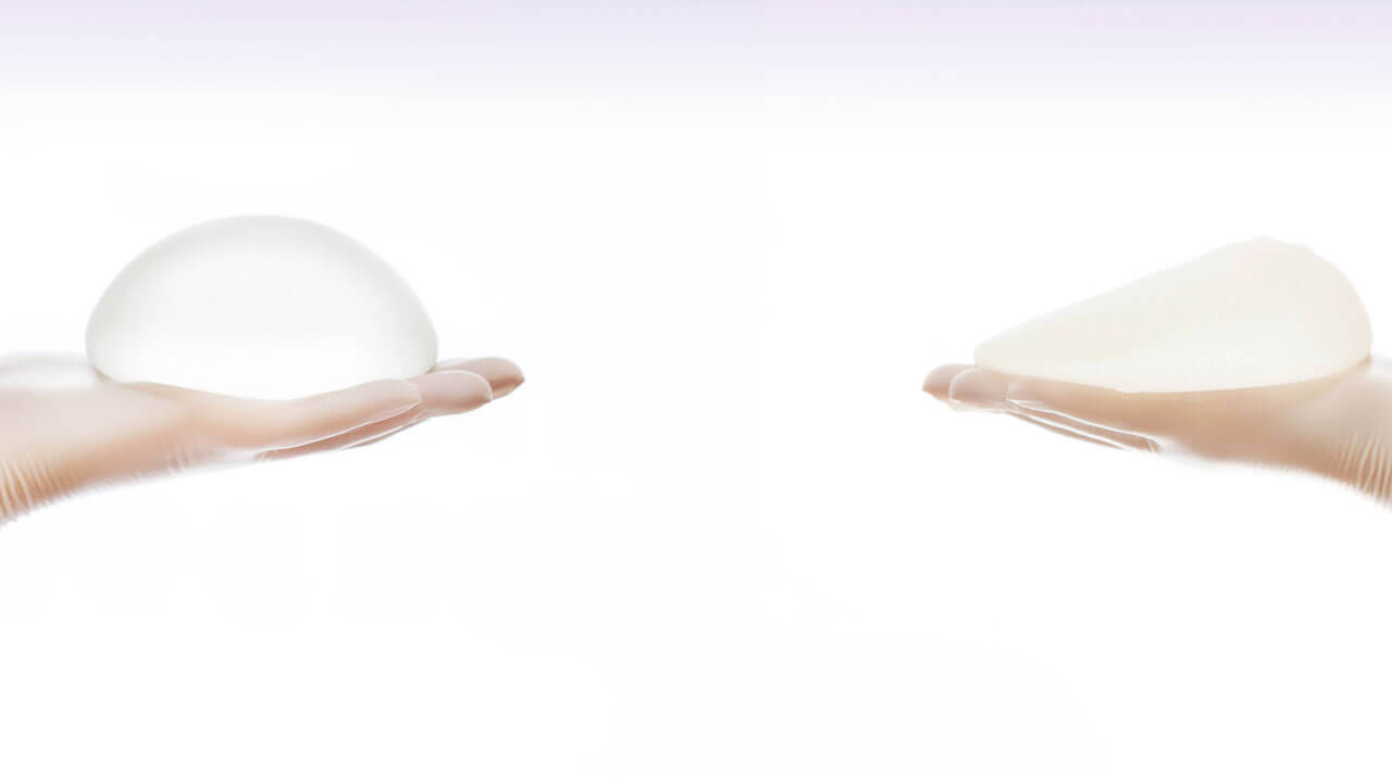 imagem demonstrando dois implantes mamários de silicone, cada um deles apoiado na palma de uma mão com luva; à esquerda um implante redondo e à direita um implante anatómico em forma de gota