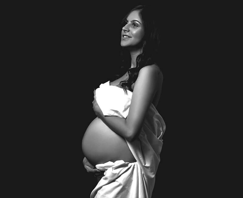 imagem lateral esquerda a preto e branco de uma mulher grávida, com a barriga à mostra e genitais e mamas cobertas por um lençol branco.