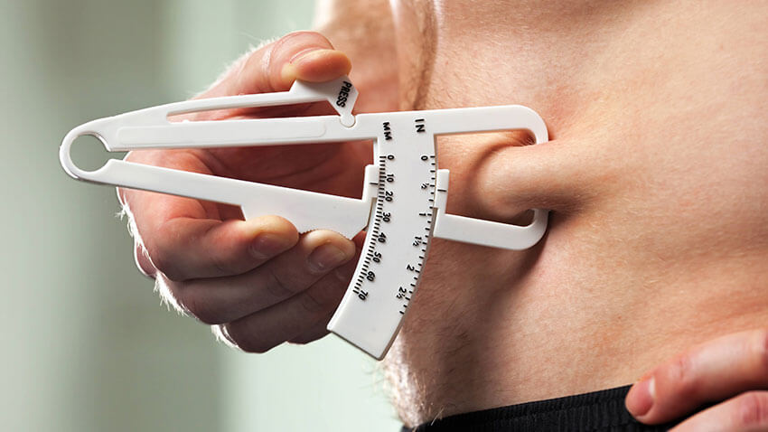 vista lateral esquerda da barriga de um homem, quantificando a gordura debaixo da pele com um calibrador, que é segurado pela sua mão direita