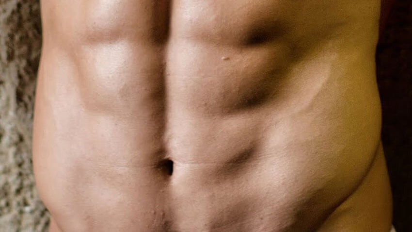 vista frontal de um homem encostado a uma parede acastanhada, demonstrando os músculos abdominais bem definidos e delineados
